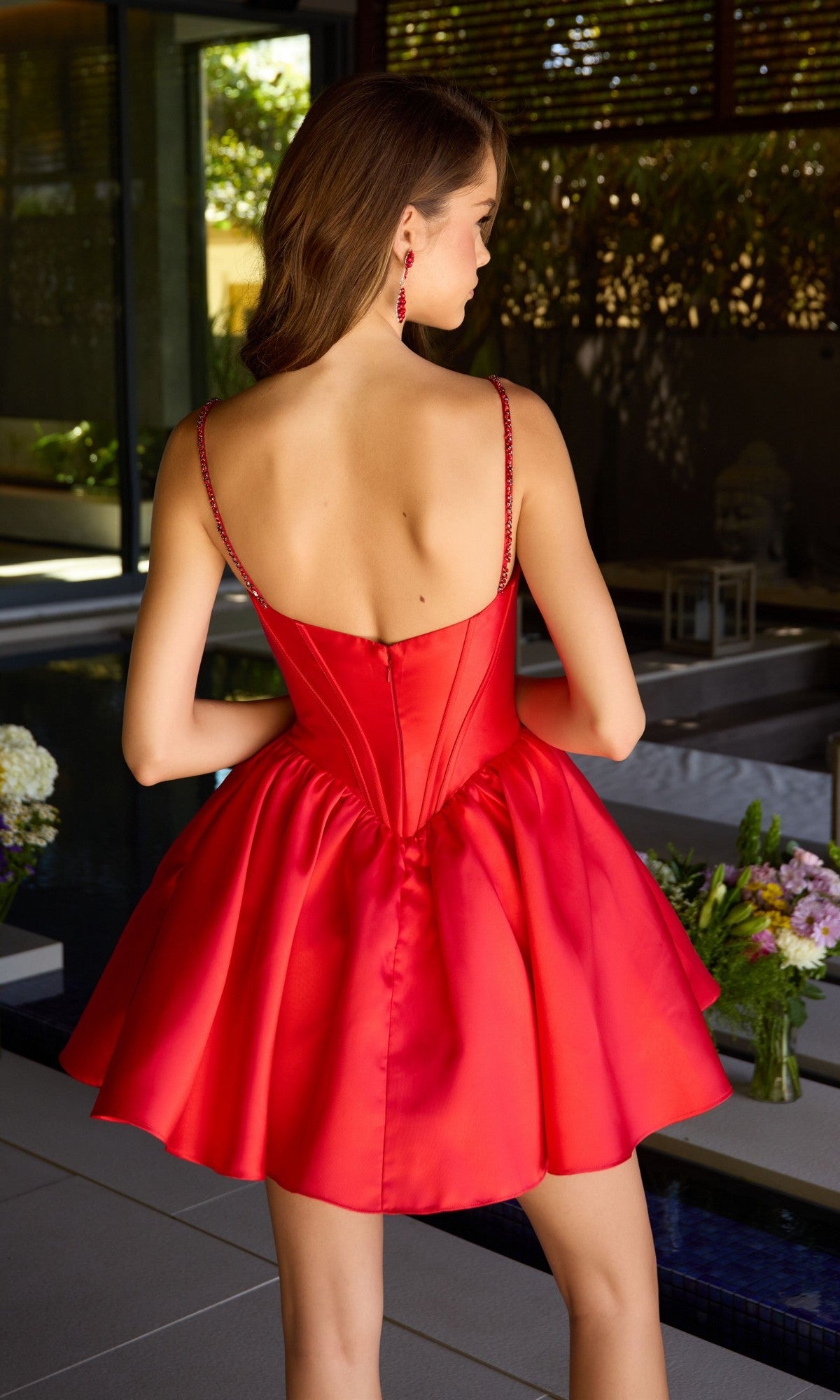 A woman stands indoors facing away from the camera, wearing the Ava Presley 42380 short red satin homecoming dress, featuring a sleeveless, backless design and flared skirt.